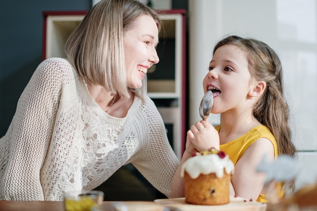 Eine Frau und ein Mädchen sitzen vor einem Kuchen, das Kind leckt einen Löffel ab.