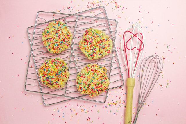 Bunte Cookies liegen auf zwei Gittern, daneben Schneebesen