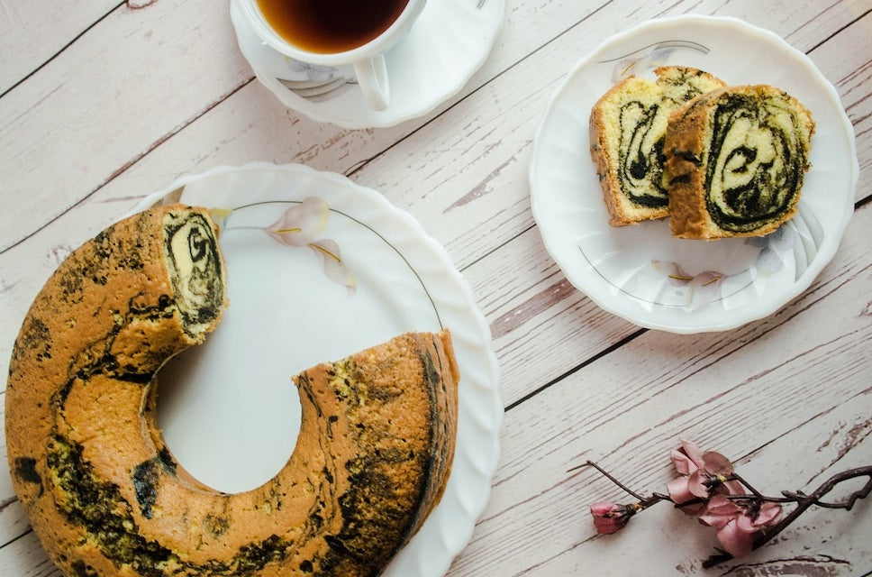Auf einem weißen Holzuntergrund steht ein großer weißer Teller mit einem angeschnittenen kranzförmigen Marmorkuchen, daneben eine Tasse Tee und ein kleiner Teller mit zwei Stücken darauf