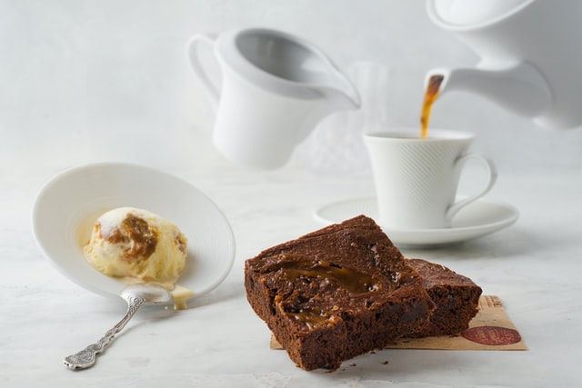 Brownies auf einem weißen Tisch. Dazu eine Kugel Eis und eine Tasse, in die Kaffee und Milch gegossen wird.