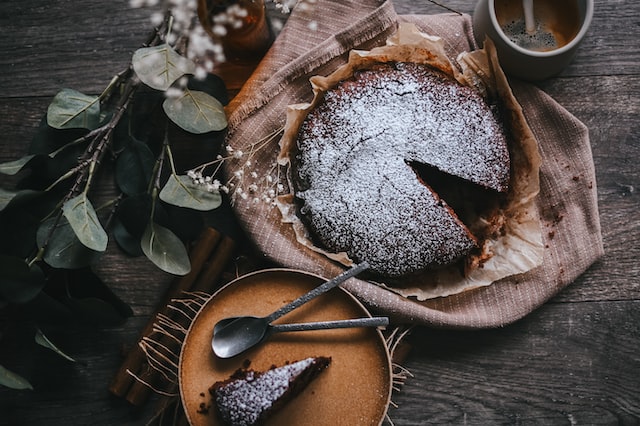 Auf einem Tisch steht ein mit Puderzucker bedeckter Schokokuchen, aus dem ein Stück rausgeschnitten wurde. Dieses liegt auf einem kleinen Teller mit Löffel daneben.