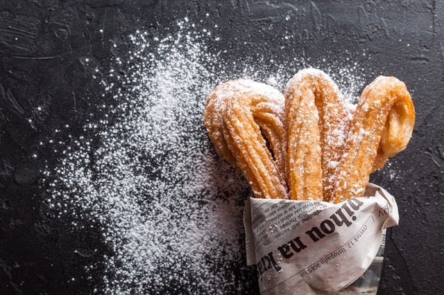 Mehrere Churros, eingewickelt in Zeitungspapier liegen auf einem schwarzen Tisch und sind mit Puderzucker bestreut.