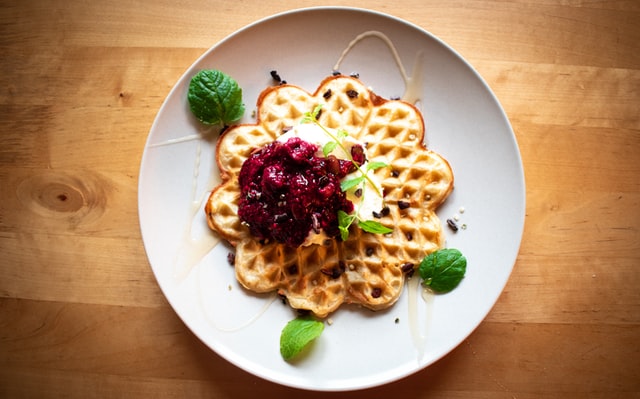 Eine Waffel auf einem Teller mit roter Grütze und Eis darauf. Drumherum sind Minzblätter drapiert.