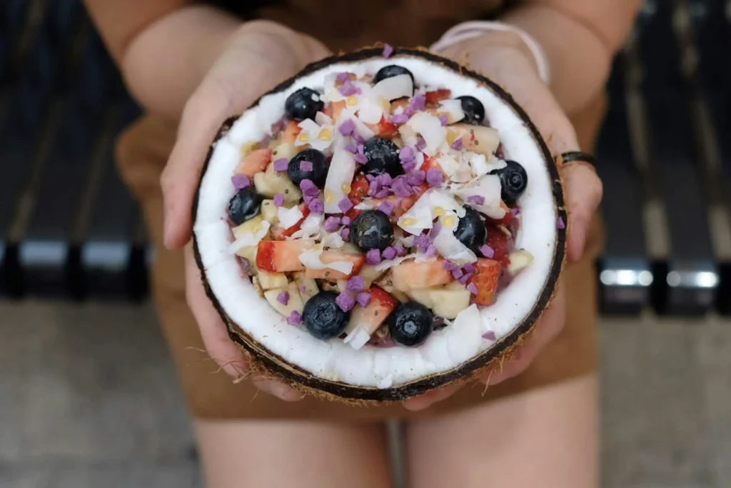 Person hält eine Kokosnussschale mit einer bunten Smoothie-Bowl, dekoriert mit frischen Früchten und Kokosraspeln.