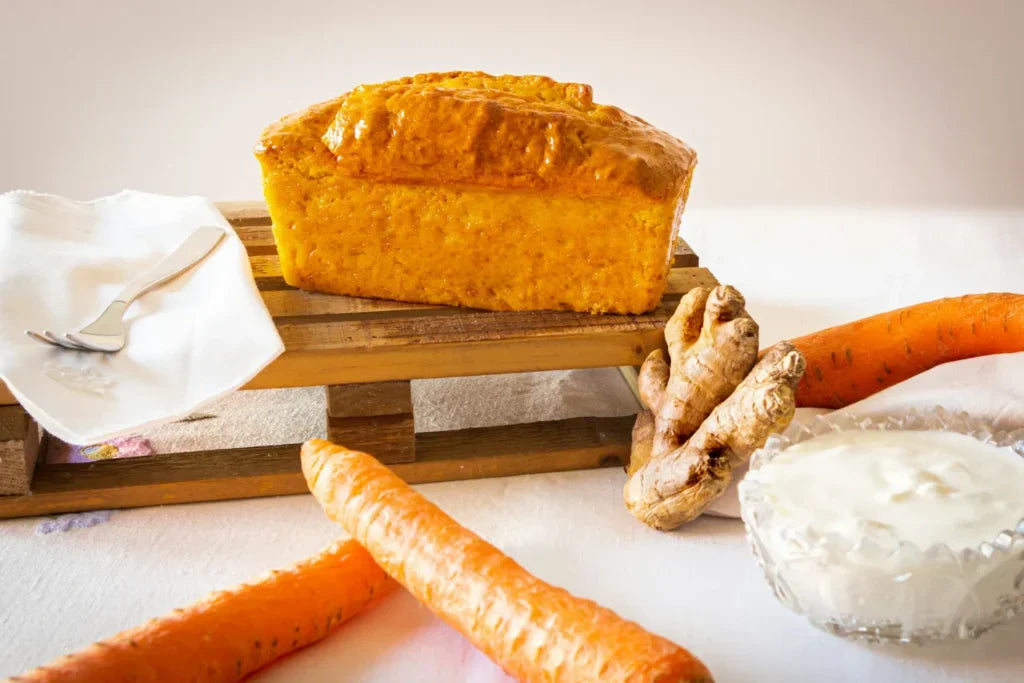 Ein goldbraunes Möhrenbrot auf einer kleinen Holzpalette, daneben frische Karotten, Ingwer und eine Schale Joghurt auf weißer Tischdecke.