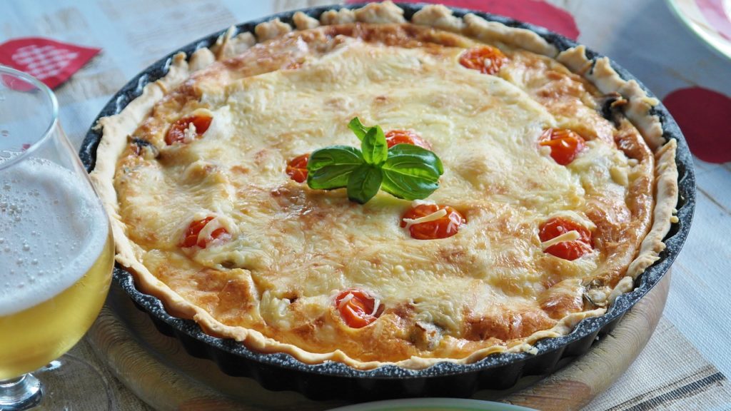 In einer Tarteform ist eine Quiche serviert, welche mit Basilikum garniert wurde. Daneben steht ein Glas mit Bier