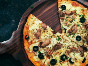 Auf einem Holzbrett ist eine Pizza angerichtet. Belegt ist sie mit Oliven, Fleisch und viel Käse.