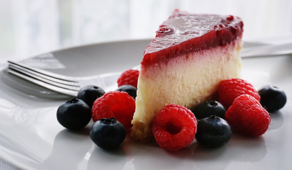 Ein Stück Käsekuchen mit einer Schicht aus rotem Fruchtpüree liegt auf einem Teller. Daneben befindet sich eine Kuchengabel und einige Him- und Blaubeeren