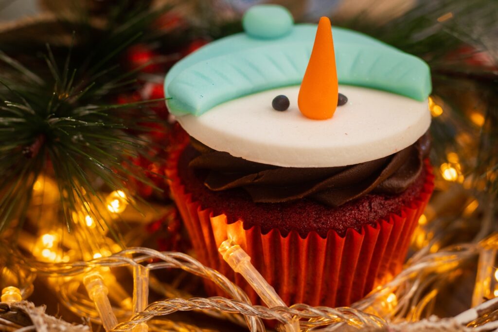 Ein Cupcake wurde mit einem Schneemann aus Fondant verziert. Um ihn liegen Lichterketten und Tannenzweige
