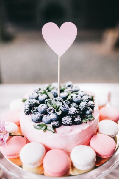Ein runder rosa Kuchen mit Blaubeeren und einem Herz als Topping ist umgeben von rosa und weißen Macarons
