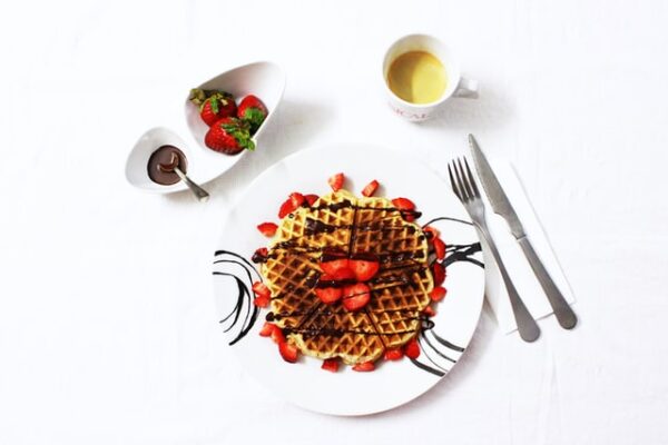 Eine Waffel, die mit Schokolade und Erdbeeren garniert ist, daneben Besteck, mehr Zutaten und eine Kaffeetasse
